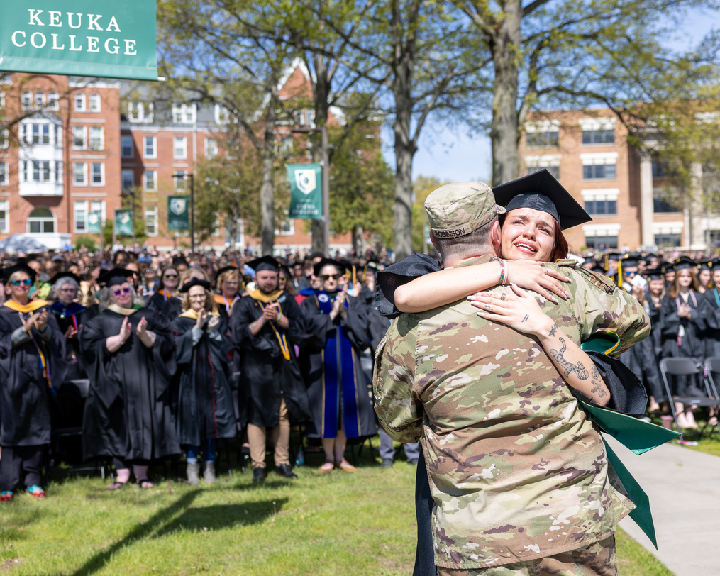 A graduating student embraces a person in military uniform in front of a large crowd during Keuka College Commencement as attendees applaud in the background.